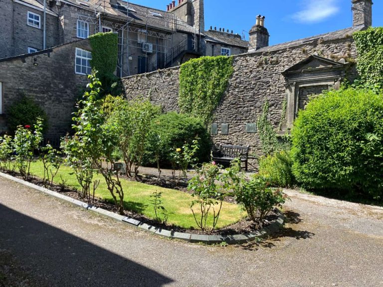 Rose garden at Kendal Unitarian Chapel in the Lake District, UK with pink flowers, green shrubs, and a wooden bench beside a winding path. This quiet garden supports the personal story about how travel feels different in retirement by showing the kind of peaceful, unexpected place that can leave a lasting emotional impression.
