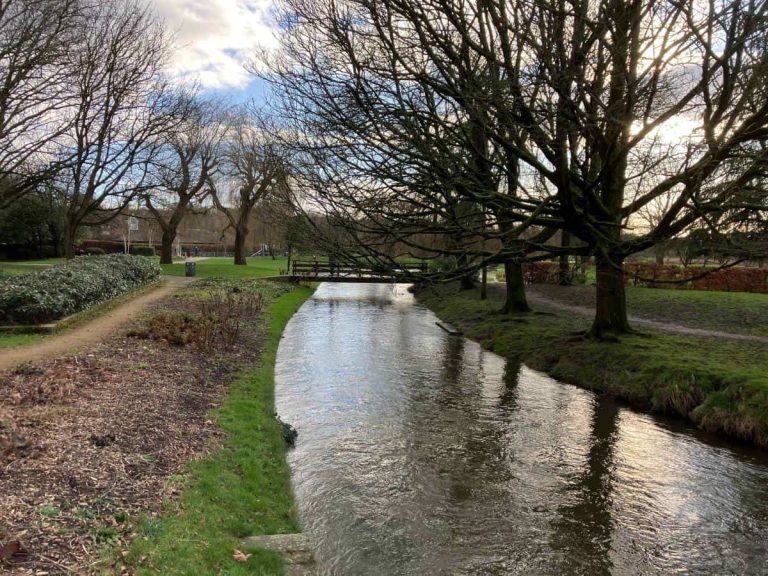 Peaceful riverside path in Salisbury, England, with a narrow stream winding through a park lined with bare winter trees and grassy banks. The quiet scene reflects the appeal of slow travel retirement, where taking time to notice local surroundings and everyday life becomes part of the journey.